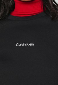 Svart sweatshirt med en ribbad röd turtleneck under; har en vit "Calvin Klein" logotyp på framsidan. Mjuk bomullsmaterial.