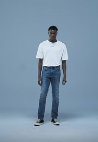 T-shirt en coton blanc et jeans en denim bleu légèrement usés. Baskets noires, chaîne argentée et une bague à la main. Fond léger uni.
