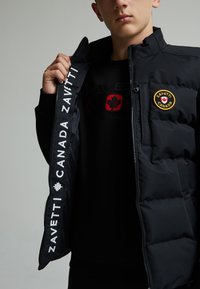 Giacca a vento nera con zip frontale, dotata di un logo circolare e scritte bianche. Tessuto liscio, design isolato e colletto alto.