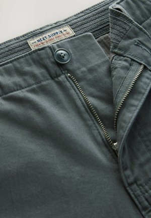 Sive pamučne hlače s metalnim patentnim zatvaračem, kopčanjem na dugme i oznakom ušivenom na pojasu. Tkanina je glatka s suptilnom teksturom.