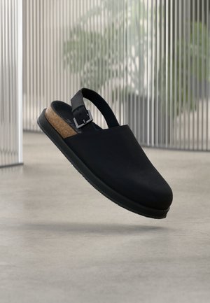 Zueco negro tipo slip-on con correa ajustable y plantilla de corcho flotando sobre suelo de hormigón, fondo con paneles verticales translúcidos.