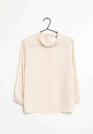 Blouse beige à manches longues avec un col rond, suspendue sur un cintre noir sur fond blanc.