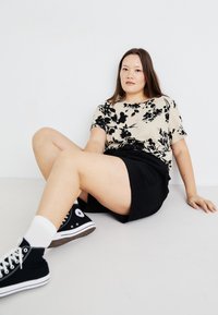 Blusa beige con estampado floral y mangas cortas, falda negra y zapatillas negras con suelas blancas. Postura sentada sobre un fondo claro.