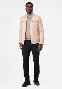 Mand iført en beige læderjakke, lys brun turtleneck-sweater, sorte bukser og sorte snørestøvler, står op mod en hvid baggrund.