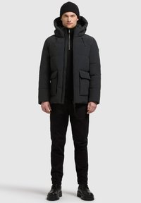 Schwarze isolierte Pufferjacke mit Kapuze, mit einem Reißverschluss, zwei Fronttaschen und einer strukturierten Oberfläche, kombiniert mit schwarzen Hosen und Stiefeln.