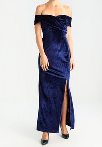 Marinblå sammetsklänning med off-shoulder-design, figursydd överdel, draperade detaljer och en hög slits vid låret. Avslutad med en lång, flödande silhuett.