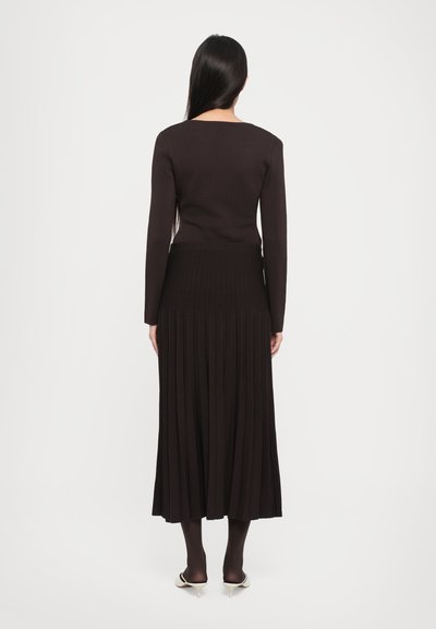 Long black pleated skirt with a fitted black top, featuring long sleeves and a square neckline, paired with black tights and heeled mules.