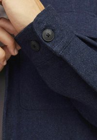 Manica di giacca in tessuto blu navy con lavorazione a trama, dotata di due chiusure con bottoni neri e un polsino piegato con cura.