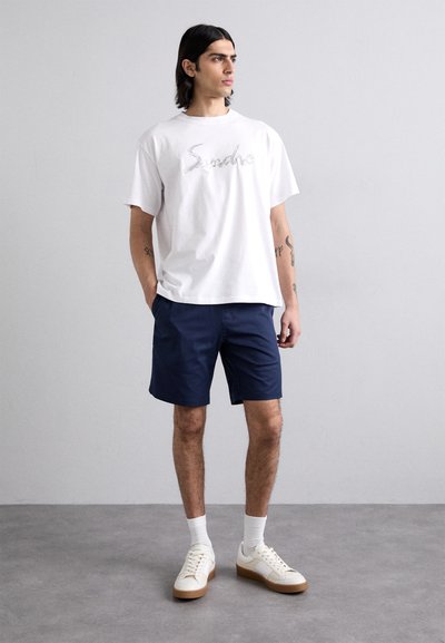 Bílý bavlněný tričko s grafikou "Sancho", spárované s námořnickými šortkami. Model má na sobě bílé tenisky a ponožky do délky crew. Minimalistický design.