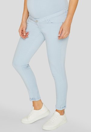 Personne enceinte portant un jean de maternité bleu clair et des baskets blanches, debout devant un fond clair uni.