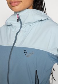 Lichtblauwe jas met een contrasterend donkerblauw onderste gedeelte, ritssluiting, verstelbare capuchon en een zwart logo op de borst.