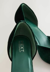 O pereche de balerini din catifea verde închis, cu vârful ascuțit, având imprimat pe branțuri "NEXT Forever Comfort", pe un fundal neutru.