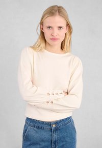 Maglione beige chiaro con texture a coste, scollo rotondo e maniche a palloncino. Accenti ritagliati notevoli vicino ai polsini. Abbinato a jeans a vita alta.