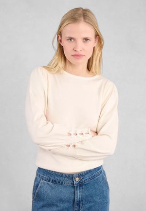 Maglione beige chiaro con texture a coste, scollo rotondo e maniche a palloncino. Accenti ritagliati notevoli vicino ai polsini. Abbinato a jeans a vita alta.