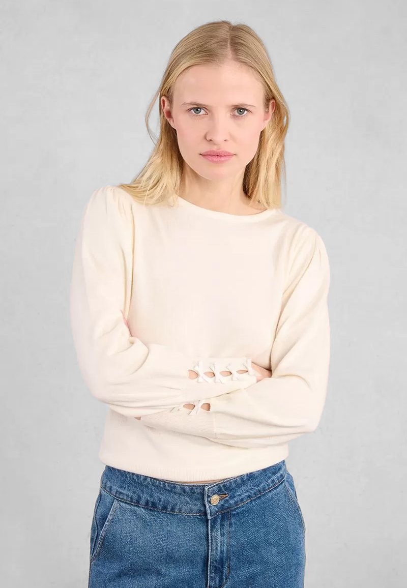 Maglione beige chiaro con texture a coste, scollo rotondo e maniche a palloncino. Accenti ritagliati notevoli vicino ai polsini. Abbinato a jeans a vita alta.