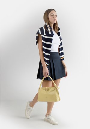 Donna con maglione a righe sulle spalle, top bianco, gonna blu navy, sneakers bianche, in mano una borsa gialla, in piedi davanti a uno sfondo semplice.