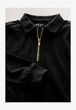 Cămașă neagră cu fermoar, realizată dintr-un material texturat, cu un fermoar auriu și guler cu rever. Manșete plisate și etichetă de marcă în interiorul gulerului.