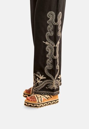 Pantalones negros con bordado floral blanco combinados con sandalias de plataforma beige con patrones geométricos negros y tiras cruzadas.