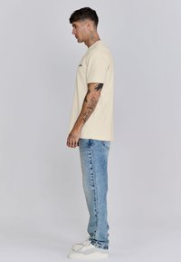 Camiseta beige de manga corta con un corte relajado, combinada con unos jeans de mezclilla azul claro y zapatillas blancas, mostrando tatuajes visibles en los brazos.