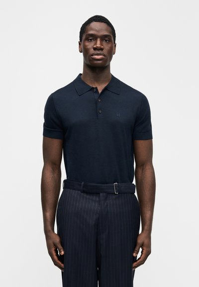 Μπλε σκούρο πόλο μπλουζάκι με κοντό μανίκι, γιακά και πατιλέτα με κουμπιά, συνδυασμένο με παντελόνι με λεπτές ρίγες και ζώνη που ταιριάζει. Λείο ύφασμα.