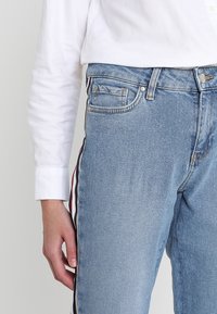 Jasnoniebieskie dżinsy o slim fit, z bocznymi paskami w kolorze czarnym, czerwonym i białym. Widoczne dodatkowe detale w postaci kieszeni i guzika.