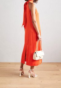 Frau trägt ein ärmelloses oranges Midikleid mit seitlichen Schlitzen, hält eine weiße gesteppte Handtasche und trägt weiße High-Heels.