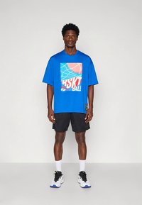 Mėlynas oversize stiliaus marškinėlis su grafiniu atspaudu ir juodi sportiniai šortai, derinami su baltais kojinėmis ir juodais sportiniais bateliais su mėlynomis detalėmis.