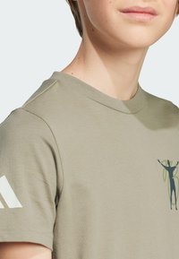 Khaki puuvillapaita, jossa on pyöreä pääntie, lyhyet hihat ja graafinen printti vihreillä yksityiskohdilla edessä.