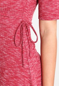 Robe rouge texturée à manches courtes, avec une taille froncée nouée par un cordon de tissu assorti. Motif subtil partout.