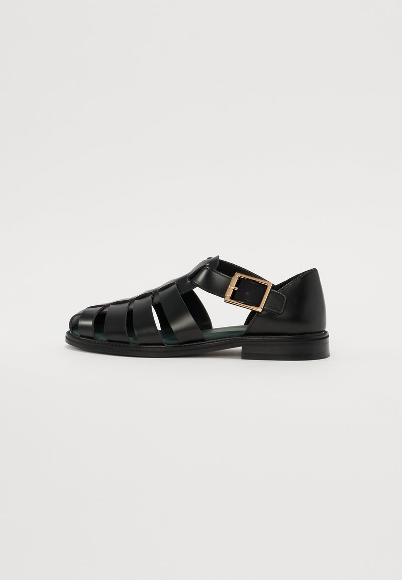 Zwarte leren sandalen met een strapontwerp, voorzien van een gouden gespaccent, open teen en een lage, platte zool. Over het geheel een gladde textuur.