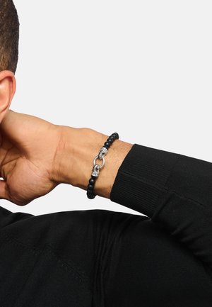 Arm eines Mannes hinter dem Kopf gebeugt, trägt ein schwarzes Perlenarmband mit zwei silbernen Schlaufenverschlüssen, gekleidet in einem schwarzen Langarmshirt.