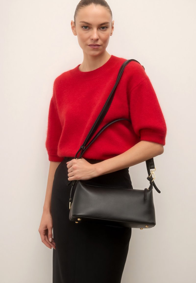 Femme portant un pull rouge à manches courtes et une jupe noire, tenant un sac à main en cuir noir avec une bandoulière.