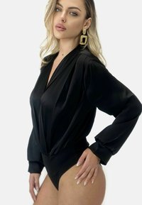 Svart bodysuit i slät tyg med V-ringning, långa ärmar och åtstramad midja; matchad med stora guldörhängen och neutral nagellack.