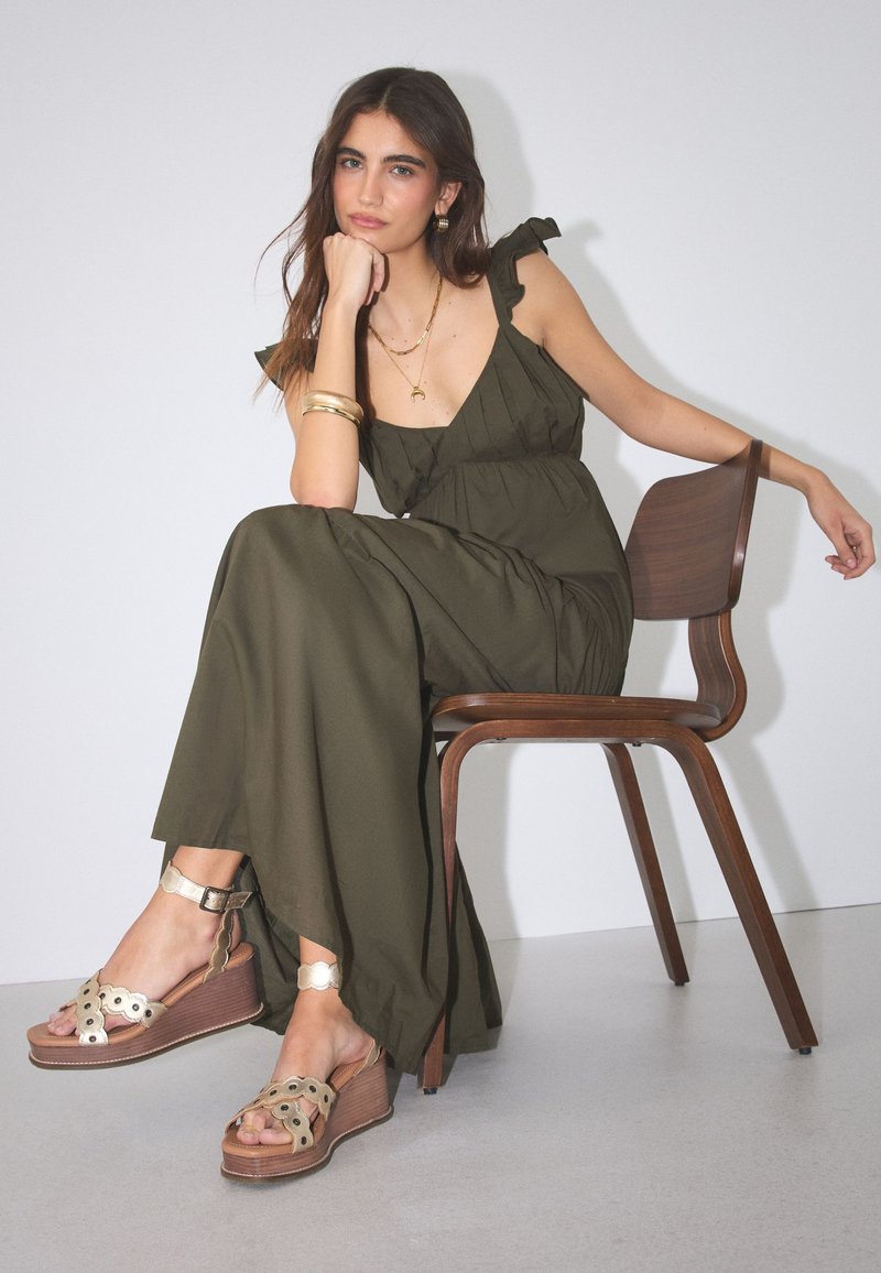 Jeune femme en robe longue sans manches vert olive et sandales compensées beiges assise de manière décontractée sur une chaise en bois contre un mur blanc.