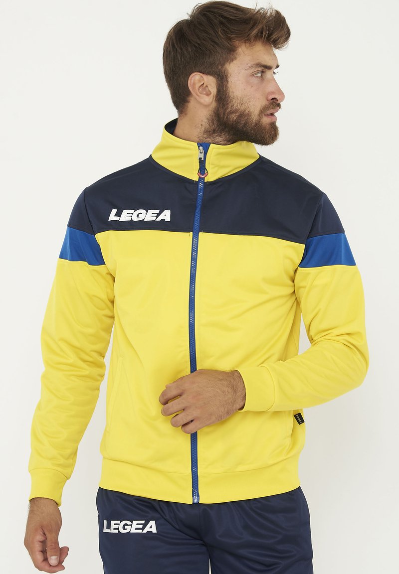 Chaqueta deportiva en amarillo y azul marino con cuello alto, acentos azules, cremallera de longitud completa y logo "LEGEA" en el pecho. Textura suave.