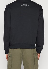 Homme portant un sweat-shirt noir avec l'inscription « Property of Gap Athletic Dept. » sur le haut du dos et un pantalon cargo vert olive, vu de dos.