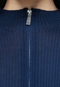 Tengerészkék bordázott pulóver magas nyakkal és ezüst színű fém cipzárral az elején, texturált kötött minta és szoros szabás jellemzi.