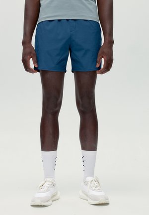 Mann trägt blaue Sportshorts, weiße knielange Socken mit Logo und weiße Turnschuhe, steht vor einem schlichten hellen Hintergrund.