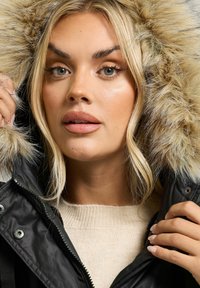 Svart parkas med fuskpäls på huva, med dragkedja och tryckknappar, bärs över en beige tröja med ribbad textur; nära bild av modedetaljer.