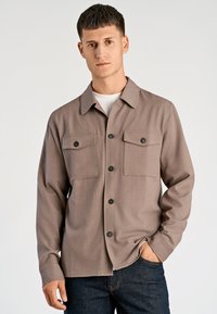 Bruin overhemd met knopen en een kraag, twee borstzakken en zwarte knopen. Gemaakt van een gestructureerde stof, gecombineerd met donkere spijkerbroeken.
