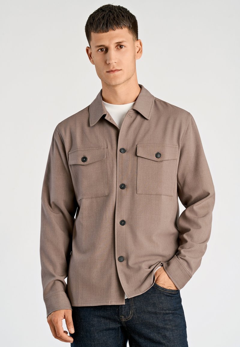 Bruin overhemd met knopen en een kraag, twee borstzakken en zwarte knopen. Gemaakt van een gestructureerde stof, gecombineerd met donkere spijkerbroeken.