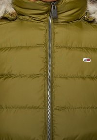 Gros plan d'une veste matelassée vert olive avec fermeture éclair centrale et petit logo rectangulaire sur le côté droit de la poitrine, dotée d'une capuche bordée de fourrure.