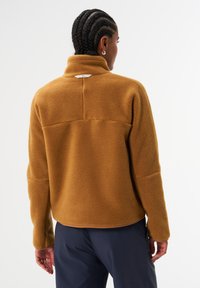 Brauner Fleece-Pullover mit hohem Kragen, der eine strukturierte Oberfläche und ein dezentes Logodetail nahe dem Hals aufweist, kontrastierend zu dunkler Hose.