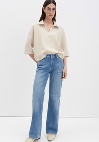 Beige top met kraag en korte mouwen, gecombineerd met lichtblauwe jeans met wijde pijpen. Model staat tegen een neutrale achtergrond. Gladde stoftextuur.