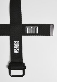 Svart tekstilrem med matte overflate, med hvit "URBAN CLASSICS" tekst og et strekkode-design merket "SPESIALUTGAVE."