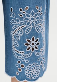 Tissu en jean bleu avec des motifs floraux découpés brodés en blanc et un bord festonné sur un vêtement.