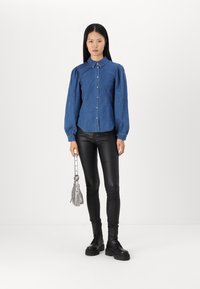 Camisa de mezclilla con botones y mangas abullonadas, color azul, combinada con pantalones ajustados negros y botas negras gruesas, sosteniendo un bolso plateado.