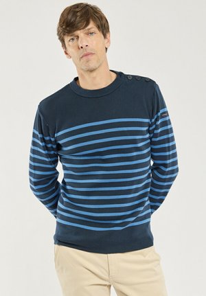 Homme portant un pull à manches longues rayé bleu marine et bleu clair avec des détails de boutons sur l'épaule, assorti à un pantalon beige, debout devant un fond uni.