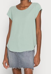 Blouse vert menthe à manches courtes avec un col rond, fabriquée en tissu léger ; associée à une jupe mini en denim gris avec des bords effilochés.