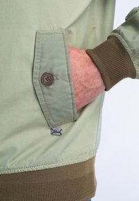 Světl zelená bunda s kapsou na knoflík, hnědým žebrovaným manžetou a malým logem. Hladká textura s prošívanými detaily.